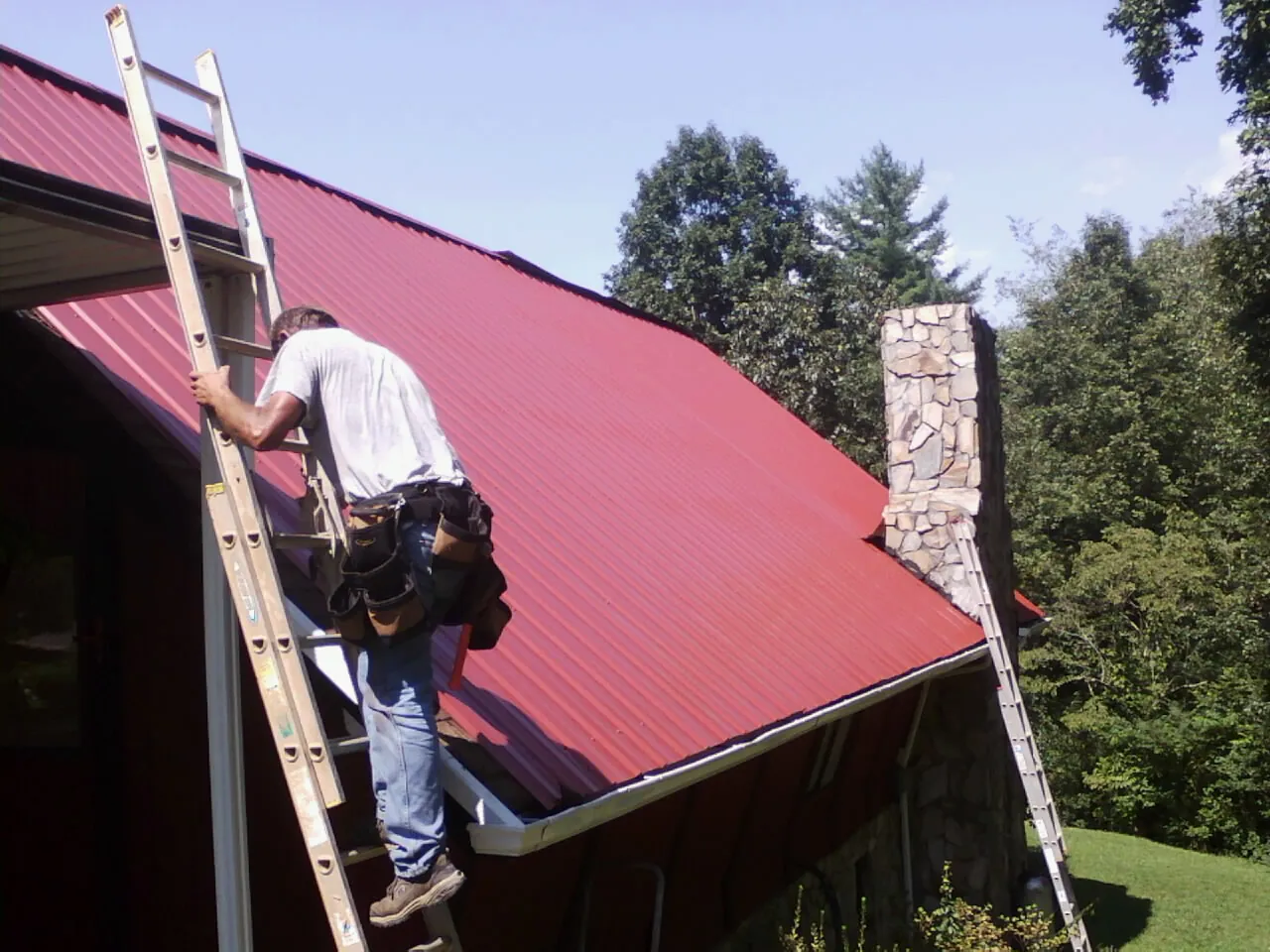 Roofer on a ladder beside a red metal roof and stone chimney — Soffit & Fascia Repair by Roofing Brothers in Tyner